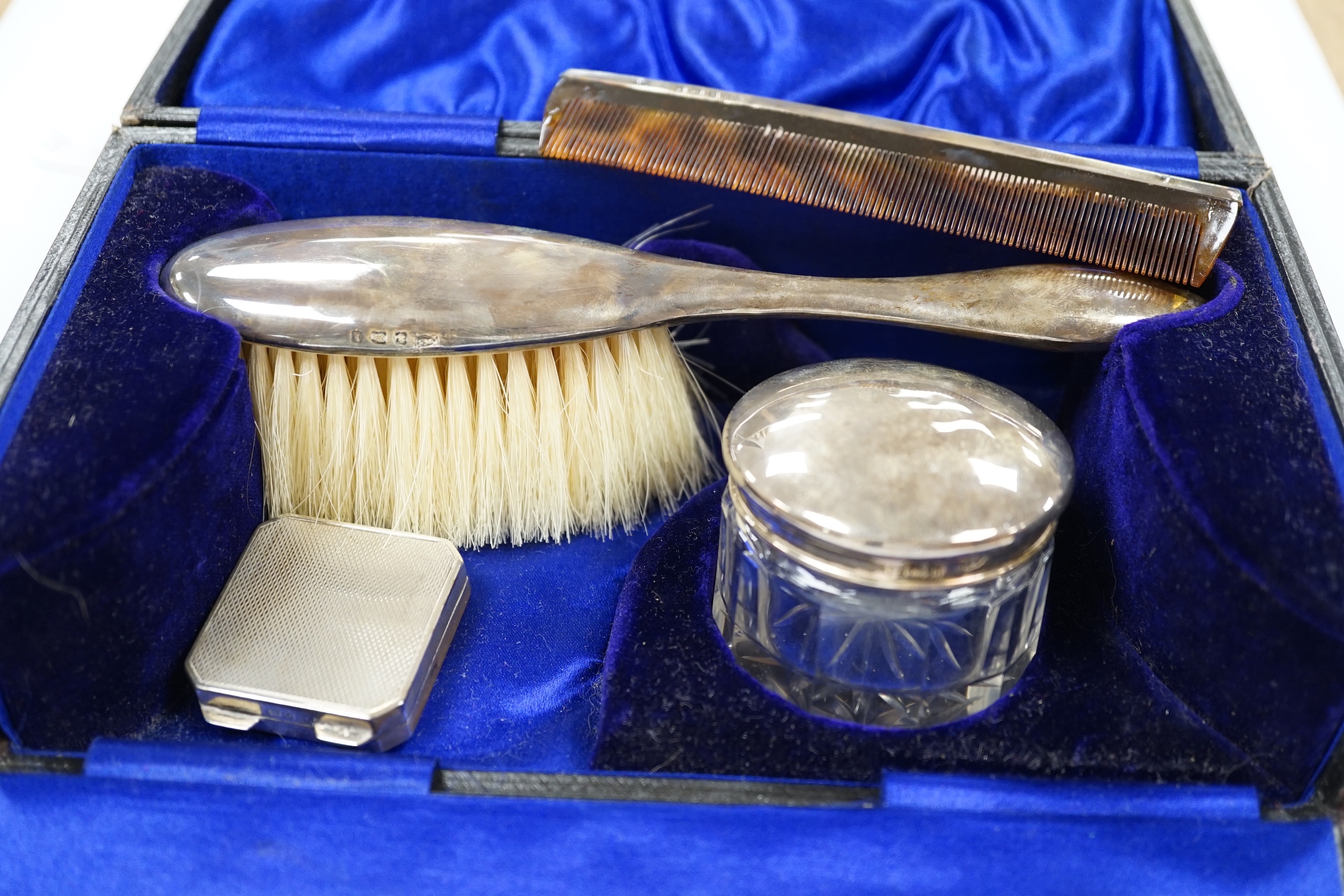 A George V engine turned silver pill box, 34mm and a cased silver mounted three piece toilet set.